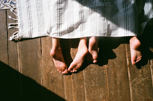 Two pairs of feet peek from under a striped blanket on a warm wooden floor, basking in natural sunlight.